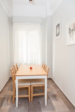 Een eetkamer met een houten tafel en stoelen, een glas rode drank, witte gordijnen en twee ingelijste foto's aan de muur.
