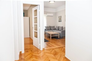 Een kamer met een houten parketvloer, een grijze bank, een klein houten bijzettafeltje en een witte deur naar een andere kamer.
