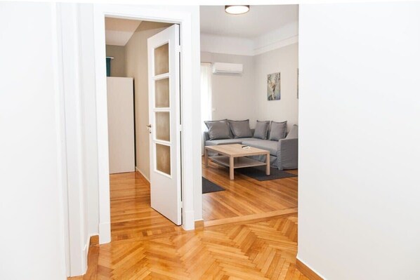 A room with a wooden parquet floor, a grey sofa, a small wooden coffee table, and a white door leading to another room.
