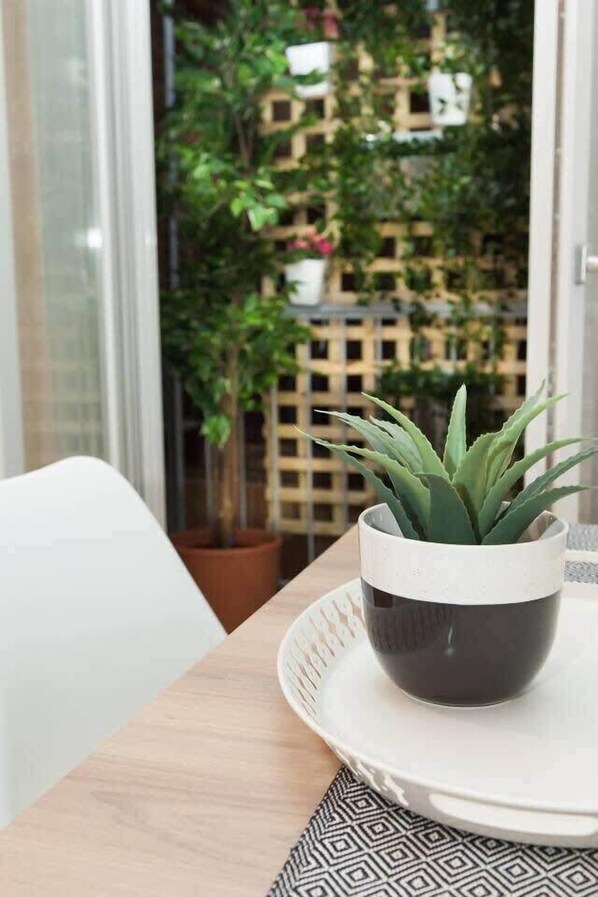 A potted plant on a table with a patterned placemat, a white chair, and a view of a building with greenery in the background.