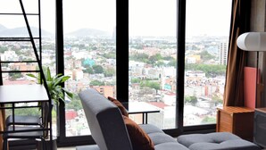A modern hotel room with a large window offering a city view, a grey sofa with a brown pillow, a small table, and a potted plant.