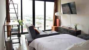 A modern hotel room with a large bed, a desk with a flat-screen TV, a small table with a potted plant, and a view of the city through large windows.