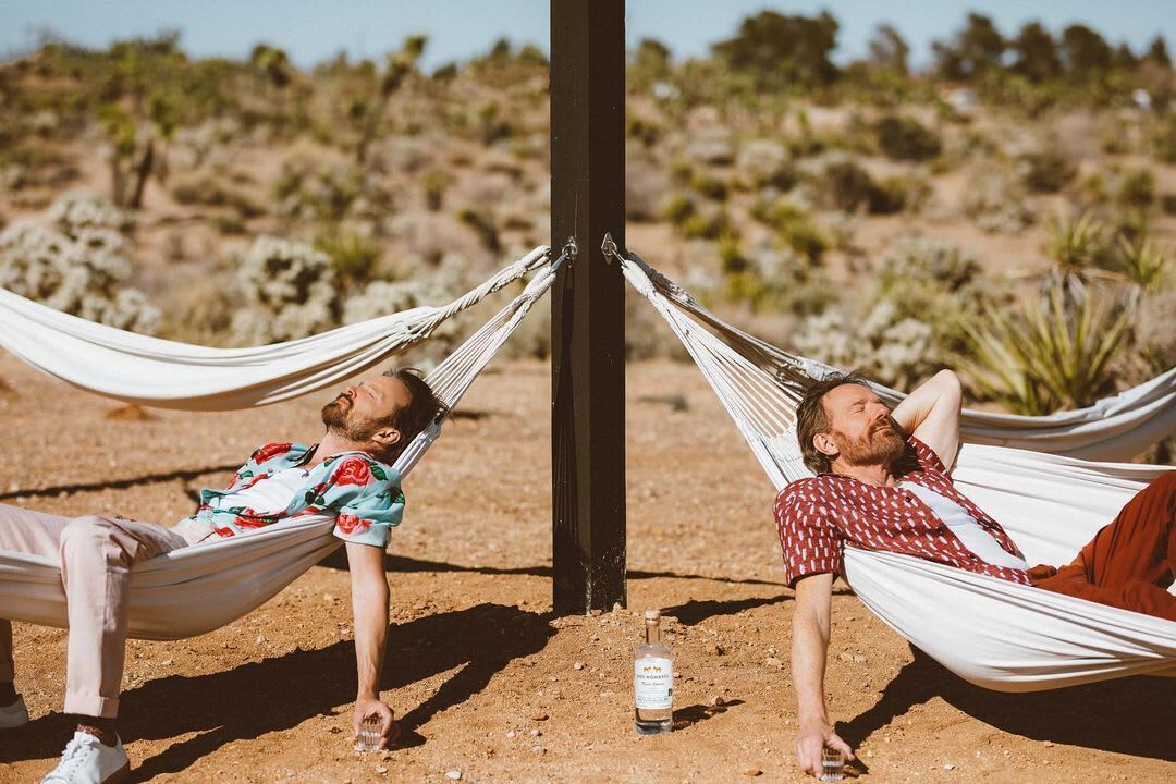 Two individuals relaxing in hammocks attached to a black pole, with a bottle of alcohol on the ground between them.