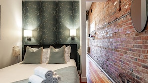 A hotel room with a brick wall, a bed with a dark headboard, and two wall-mounted lights. - Gateway Inn: Cora's Loft (Stratford)