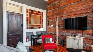 A living room with a brick wall, a fireplace, a TV mounted on the wall, a red chair, and a small table. - Gateway Inn: Cora's Loft (Stratford)