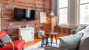 A cozy living room with a brick wall, a flat-screen TV, a radiator, and a large window. - Gateway Inn: Cora's Loft (Stratford)