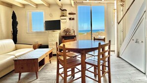 Ein gemütliches Wohnzimmer mit Esstisch und Stühlen aus Holz, einer Couch, einem Fernseher und Meerblick durch große Fenster.