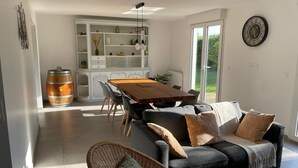 A modern living room with a dark grey sofa, a wooden dining table, and a wicker chair. There is a large window providing natural light and a view of greenery outside. - New : House, fully renovated, with garden, 200m from the beach - 3*** French property (Loctudy)