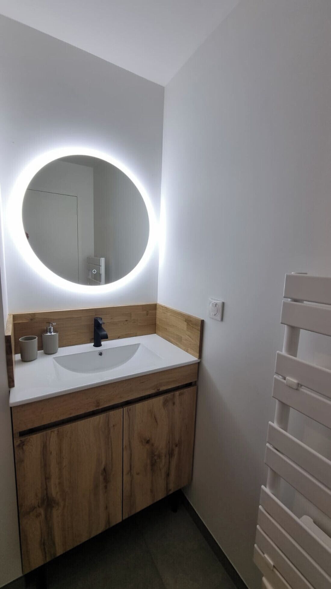 A bathroom with a white sink, wooden vanity, and a round mirror with a light.