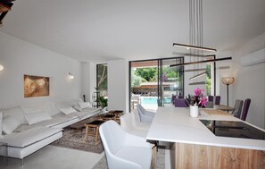 Un salon moderne avec un canapé blanc, une table à manger et des chaises, et une vue sur un espace piscine extérieur à travers des portes vitrées.
