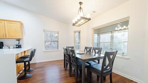 Un comedor con una mesa y sillas de madera, una encimera de cocina con taburetes y ventanas amplias con vistas a árboles.