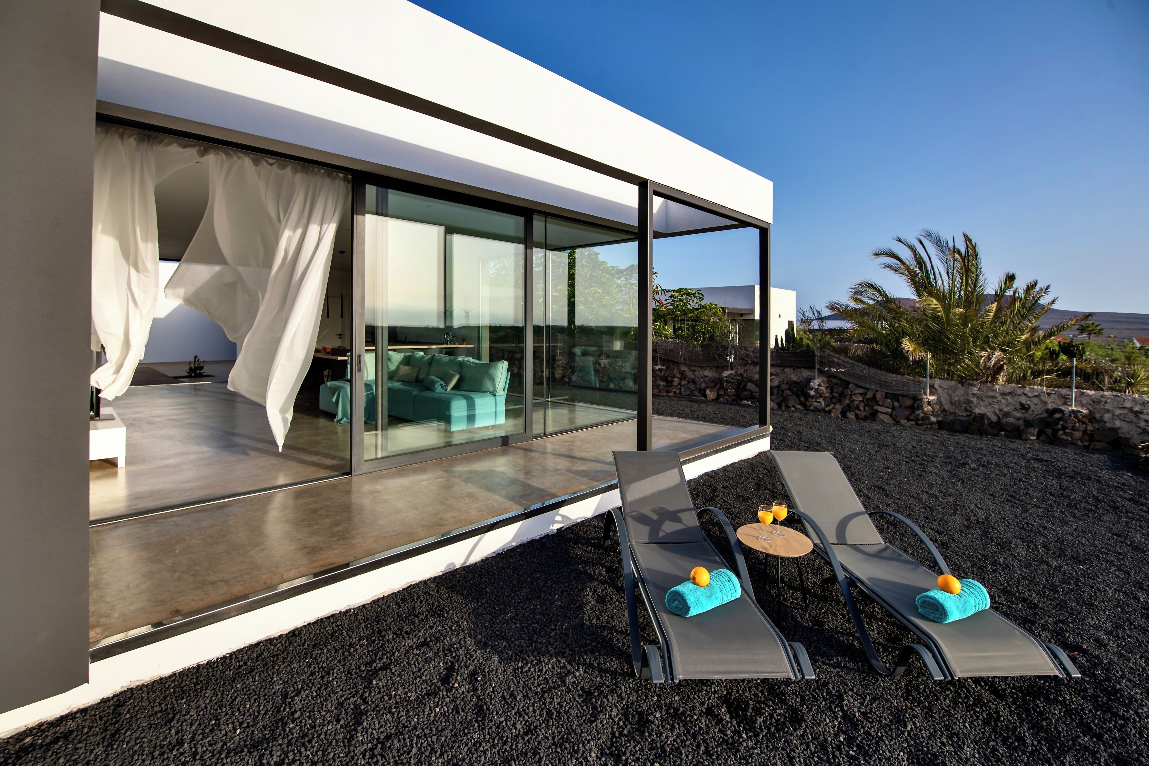 A modern outdoor lounge area with two sun loungers, a sofa, and a glass-enclosed room with a view of the landscape.