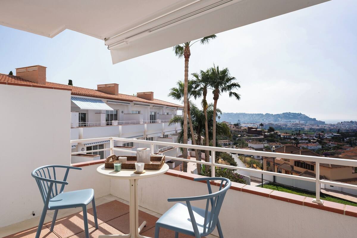 A balcony with two chairs, a table, and a view of a cityscape with palm trees.