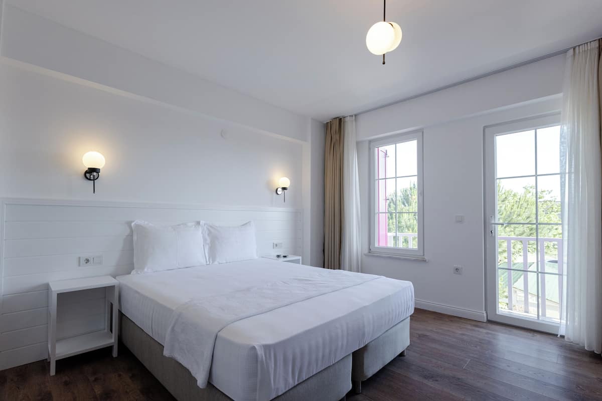 Uma cama grande e bem arrumada em um quarto claro, com duas mesas de cabeceira, uma janela ampla e uma porta que dá para área externa.