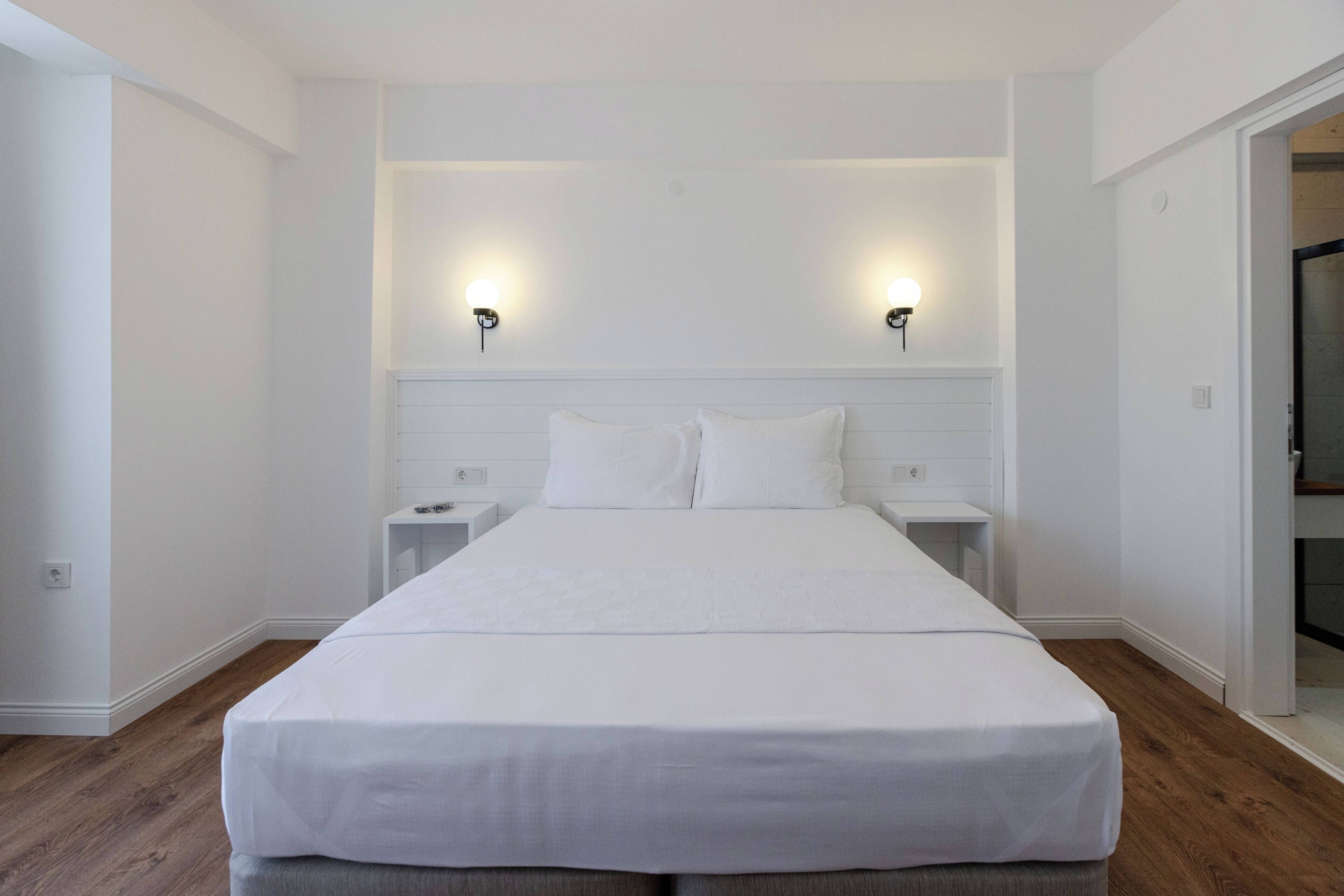 Una cama grande y bien hecha en una habitación con paredes blancas y suelo de madera. Hay dos mesitas de noche con lámparas a ambos lados de la cama.