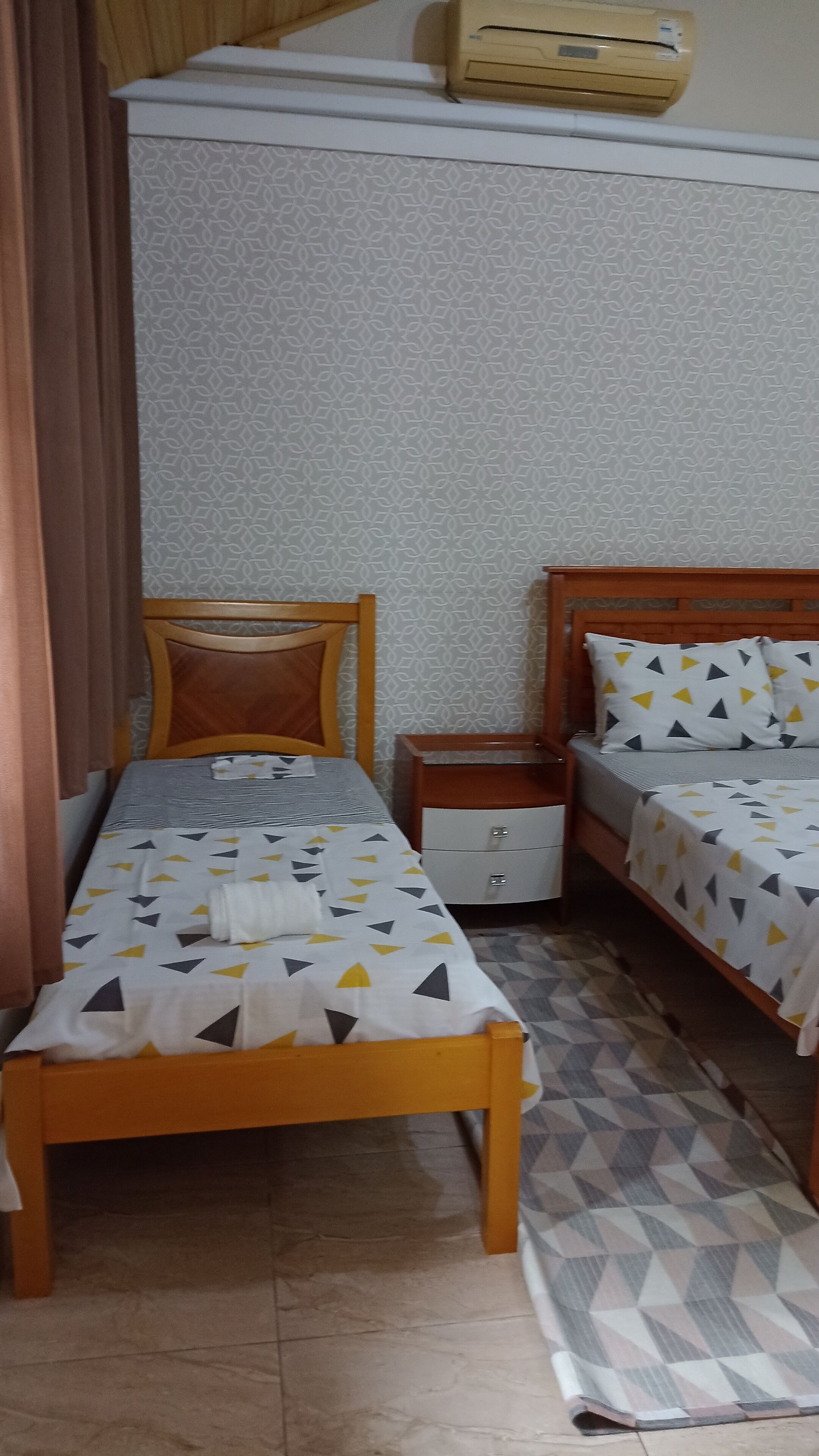 Una habitación pequeña y ordenada con dos camas individuales, una mesita de noche, un armario pequeño, papel tapiz estampado y un aire acondicionado instalado en el techo.