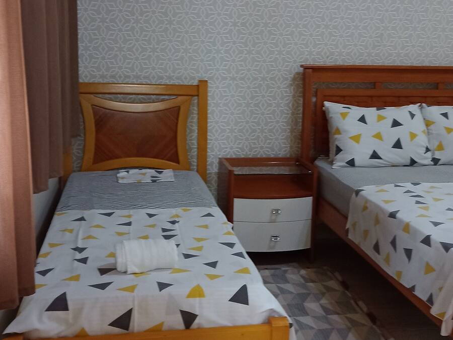 Una habitación pequeña y ordenada con dos camas individuales, una mesita de noche, un armario pequeño, papel tapiz estampado y un aire acondicionado instalado en el techo.