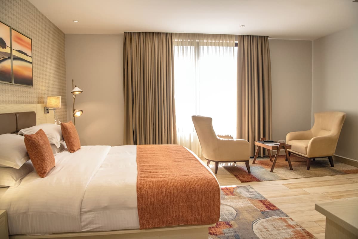 A hotel room with a large bed, two armchairs, a small table, and a view of the outdoors through the window.