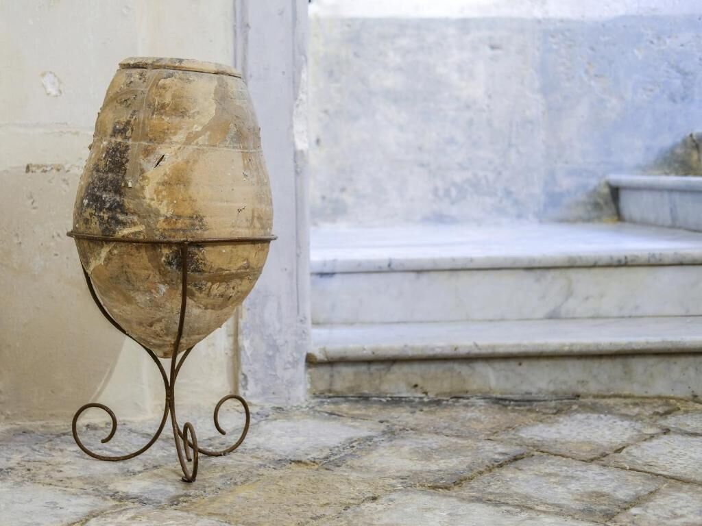 A weathered, rustic outdoor urn on a metal stand, positioned on a stone-paved area near a set of steps leading up to a building.