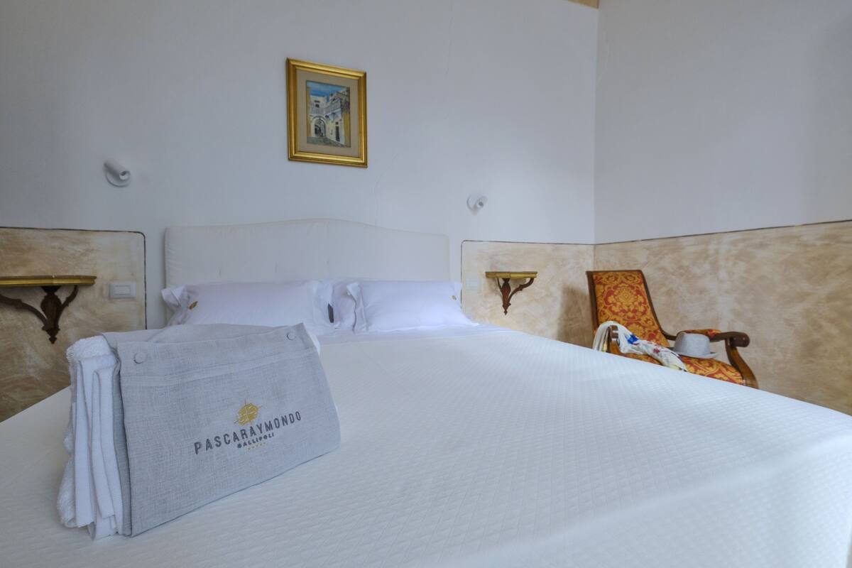 A neatly made bed with a towel and a robe on it, a chair with a patterned cushion, and a framed picture on the wall.