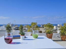 Una mesa con dos vasos de bebidas y plantas en macetas en una terraza en la azotea, con vista al mar y edificios.