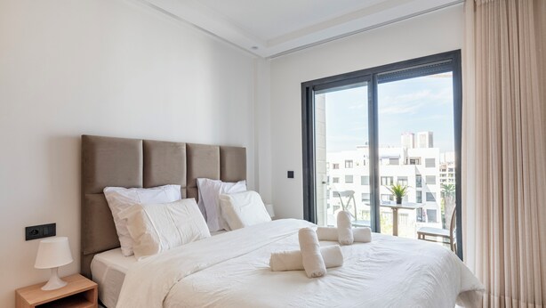 A hotel room with a large bed, a nightstand, a window with curtains, and a view of buildings outside. - Charming Apartment 42 (Casablanca)