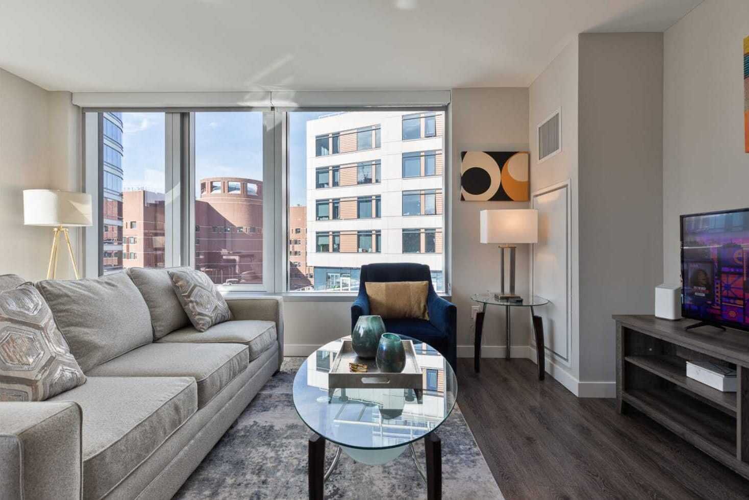 A modern living room with a sofa, a glass coffee table, a chair, a TV, and a view of buildings through large windows.