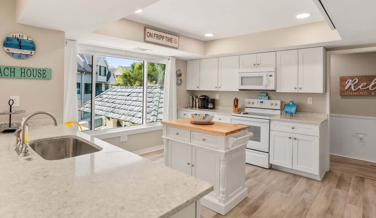 A kitchen with white cabinets, a wooden island, a sink, and a window with a view of a house.