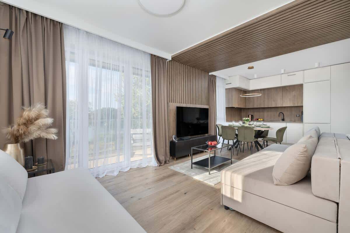 A modern living room with a sofa, a coffee table, and a TV. There is a kitchen area with cabinets and a dining table.