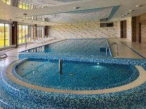 Une grande piscine intérieure avec des bords carrelés, entourée d’une plage et d’une balustrade en verre.