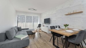 A modern living room with a grey sofa, a dining table set for four, and a TV mounted on the wall. - Stylish Apartment with Terrace | Spa Access and Parking (Dziwnów)