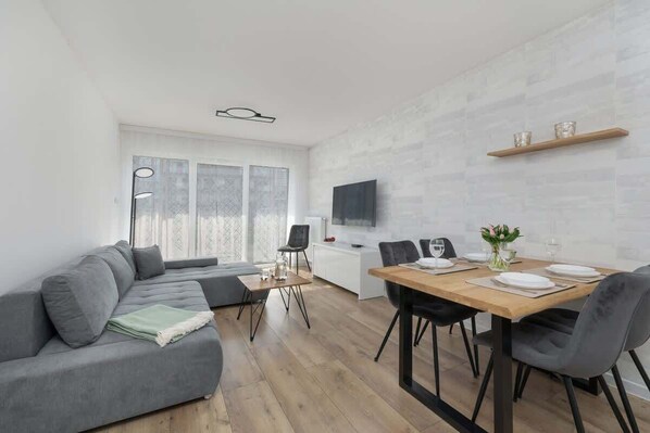 Un salon moderne avec un canapé gris, une table à manger dressée pour quatre personnes et un téléviseur fixé au mur.