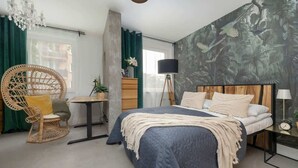 Quarto com uma cama grande, uma cadeira de vime, uma mesinha e um candeeiro de chão.