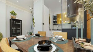 Uma área de refeições moderna com uma mesa de madeira posta para quatro pessoas, rodeada por cadeiras. Uma tigela preta e pratos brancos estão sobre a mesa, com um vaso de flores ao lado.