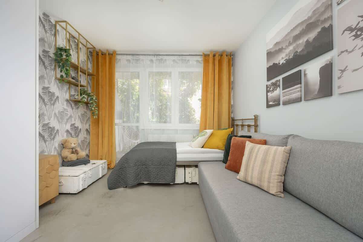 A cozy bedroom with a bed, a grey sofa, a teddy bear, and a window with curtains.