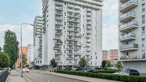 A modern high-rise residential building with multiple balconies, a paved sidewalk, and parked cars.