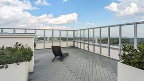 A spacious balcony with a wooden floor, a single lounge chair, and a view of a cityscape.