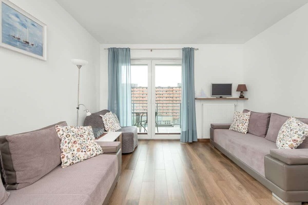 A bright, spacious living room with a comfortable sofa, soft cushions, and a large balcony window in the apartment at Obrońców Wybrzeża 15, Gdańsk.