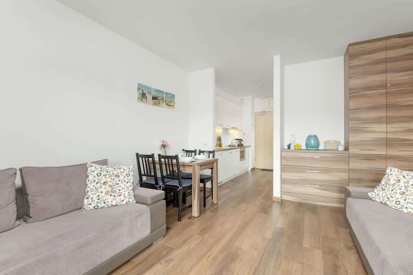 A modern living room with a sofa, a dining table with chairs, a kitchen area, and a wooden wardrobe.