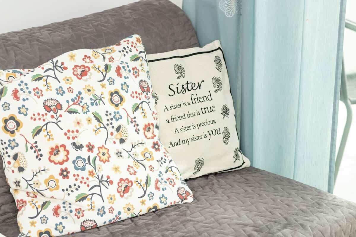 A bed with a floral pillow and a cushion with a heartfelt message about a sister.