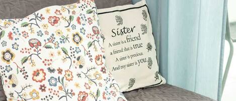 A bed with a floral pillow and a cushion with a heartfelt message about a sister.