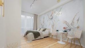 A modern bedroom with a wooden floor, a bed with a white headboard, a bedside table with a lamp, and a wall with a bird mural.