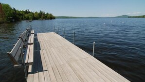 Un muelle de madera se adentra en un lago tranquilo, con cisnes a lo lejos y una orilla bordeada de bosque.