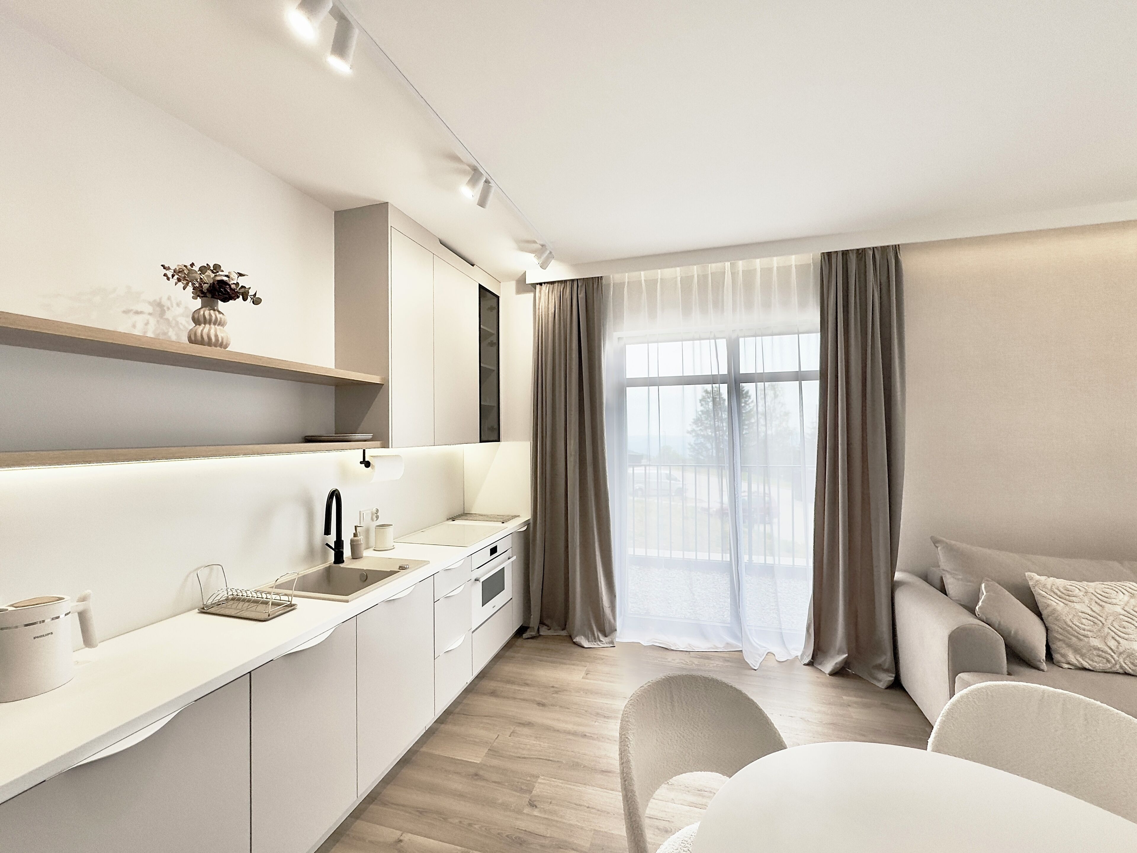 Cozinha moderna com pia embutida, armários brancos e bancada de madeira. Há uma janela com cortinas leves e vista para o exterior.