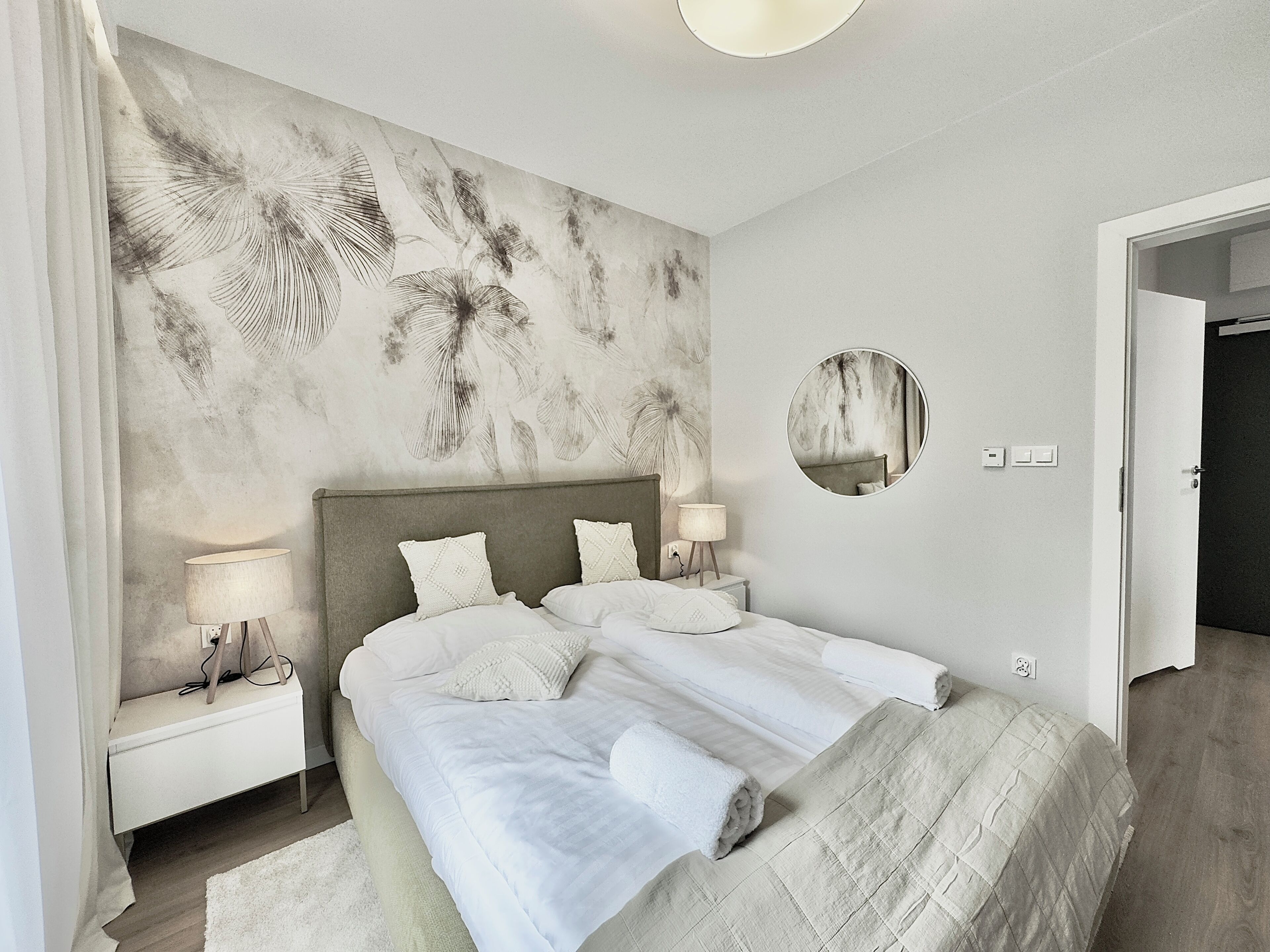 Un dormitorio con una cama grande, dos mesitas de noche, un tocador, un espejo redondo y una pared con estampado floral.