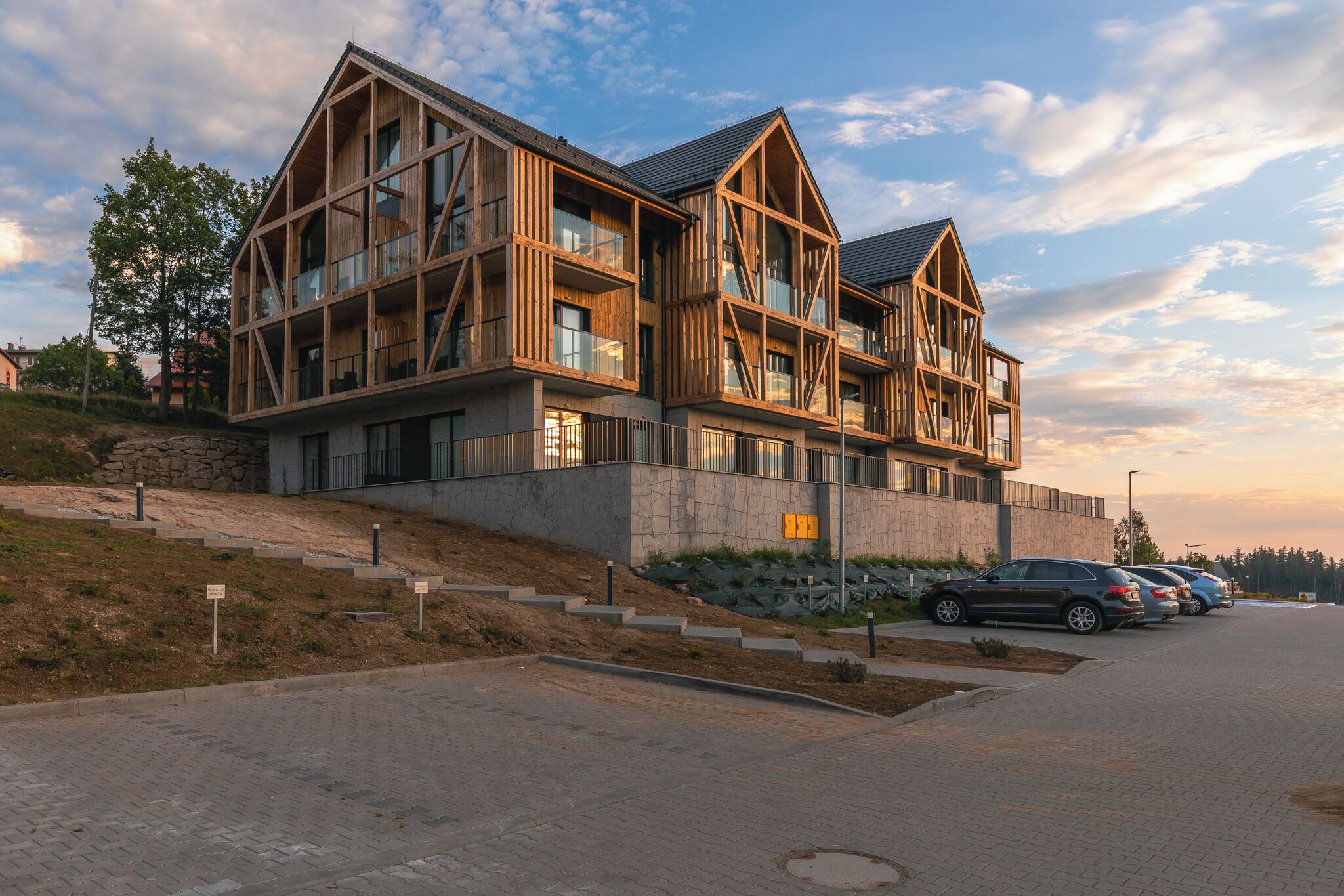 A modern multi-story building with a wooden facade and large windows, situated on a sloped site with a paved driveway and parked cars.
