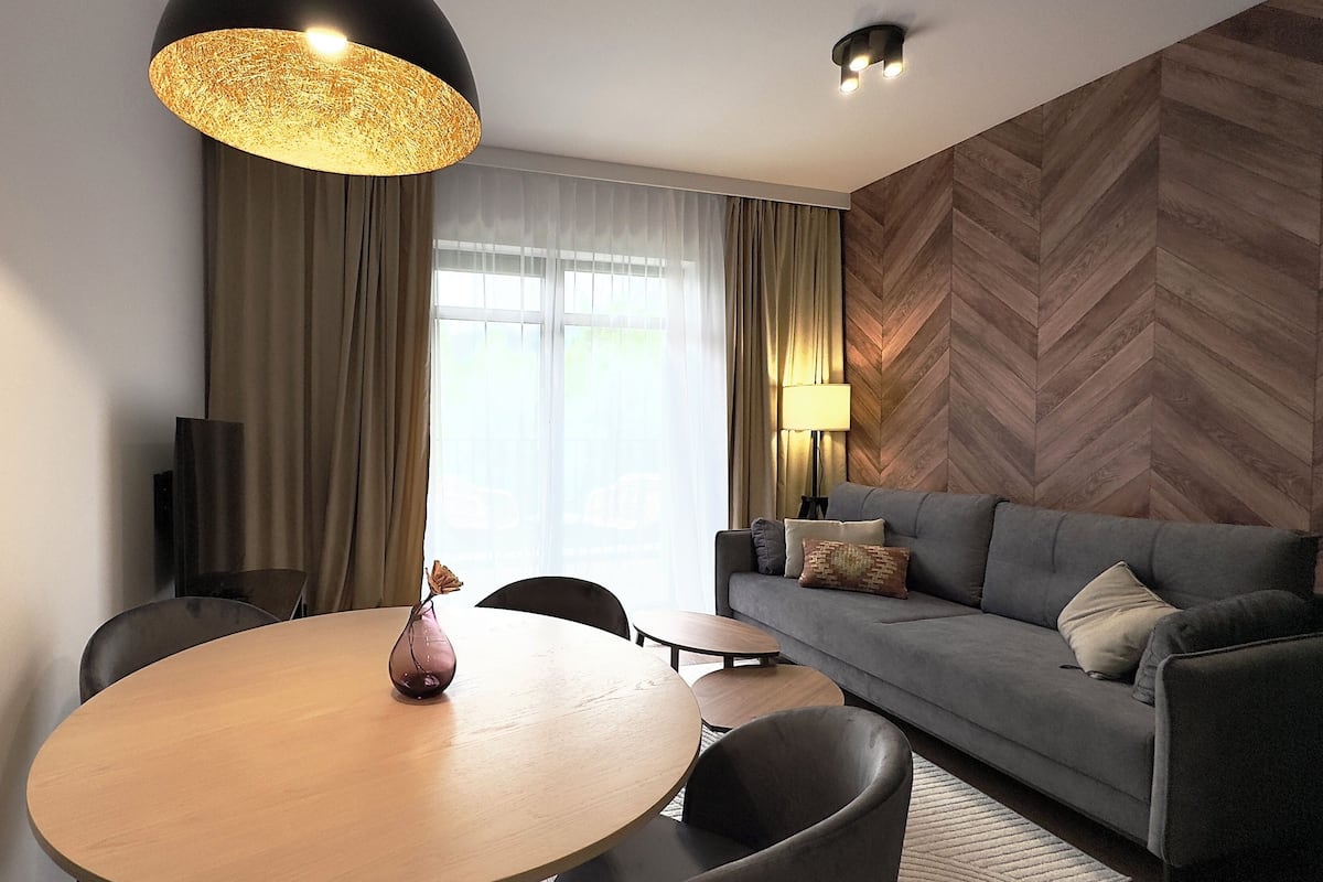 A modern hotel room with a round wooden dining table, a grey sofa, and a large window with curtains.