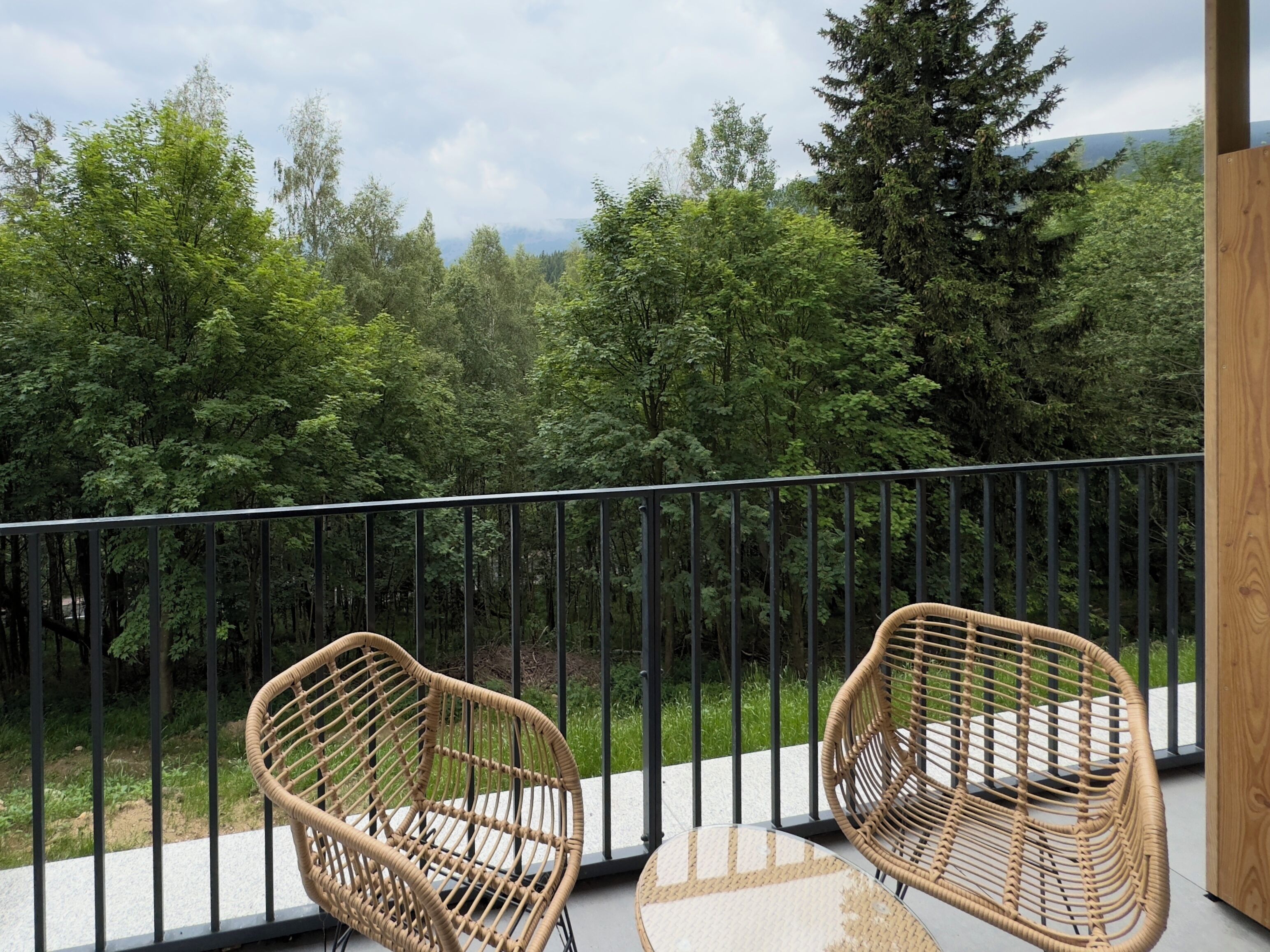 A balcony with two wicker chairs and a table, overlooking a forested area with a view of distant mountains.