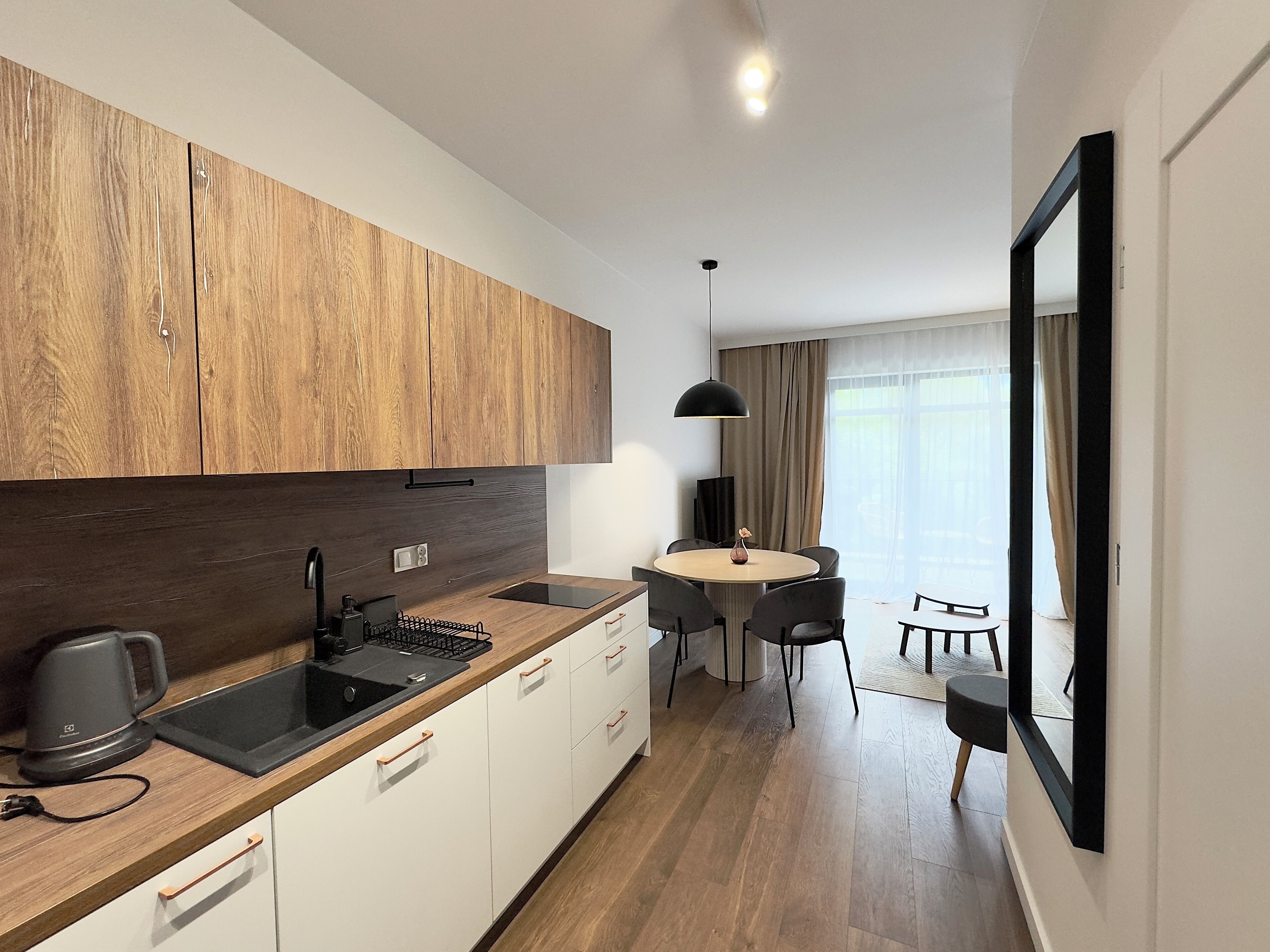 A modern kitchen with wooden cabinets, a black sink, and a kettle. There is a dining area with a table and chairs, and a large mirror on the wall.