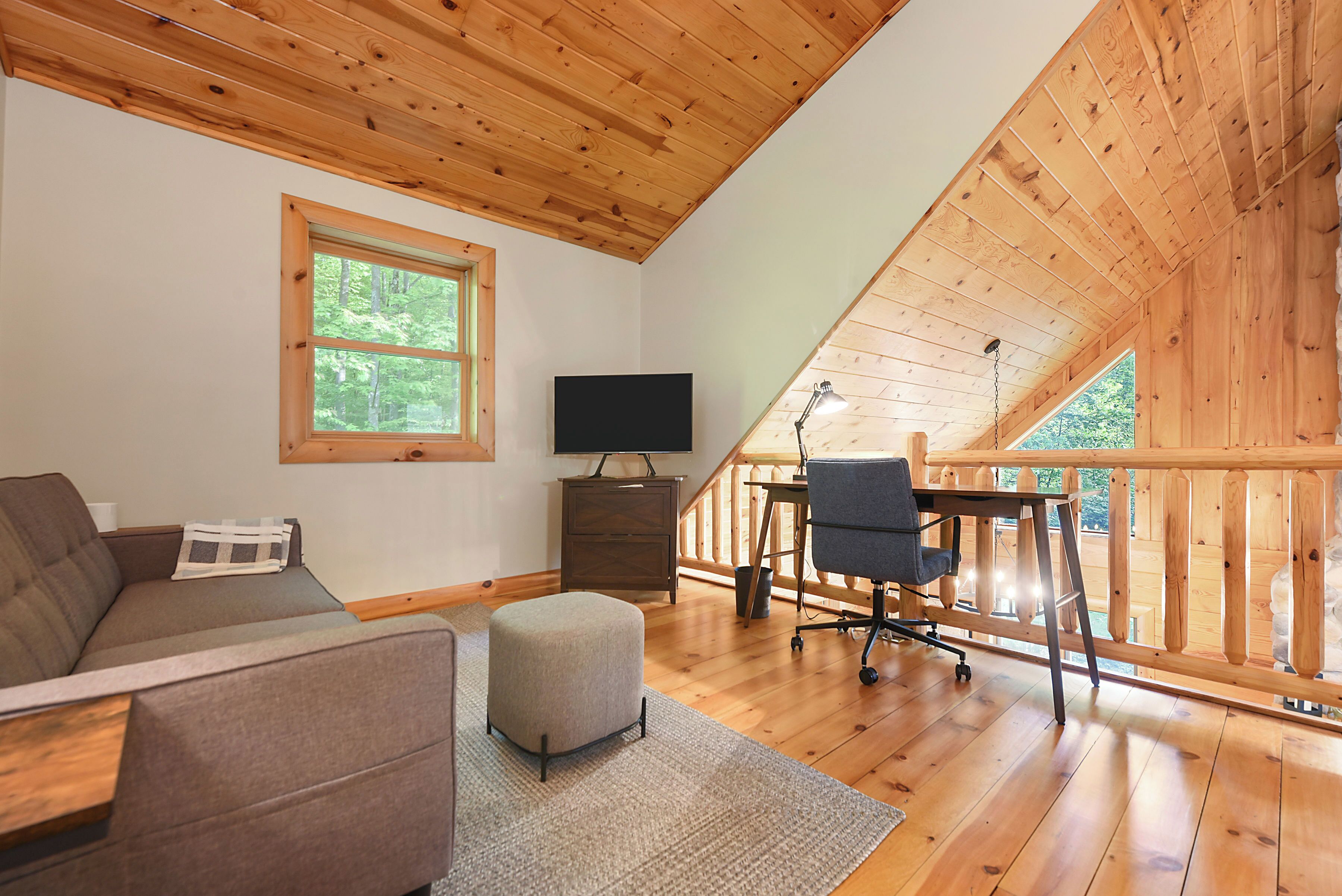 A cozy living room with a sofa, ottoman, and a desk with a chair. There's a TV on a stand and a window with a view of trees.